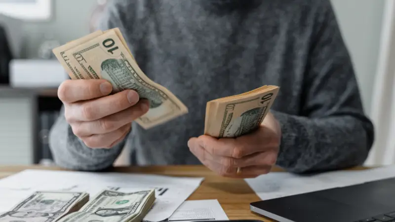 A person wearing a gray sweater is sitting at a desk, counting multiple stacks of U.S. $10 bills. The desk is cluttered with paperwork, loose bills, and a laptop, indicating financial activity or budgeting. The image conveys themes of money management, financial planning, or expense tracking.