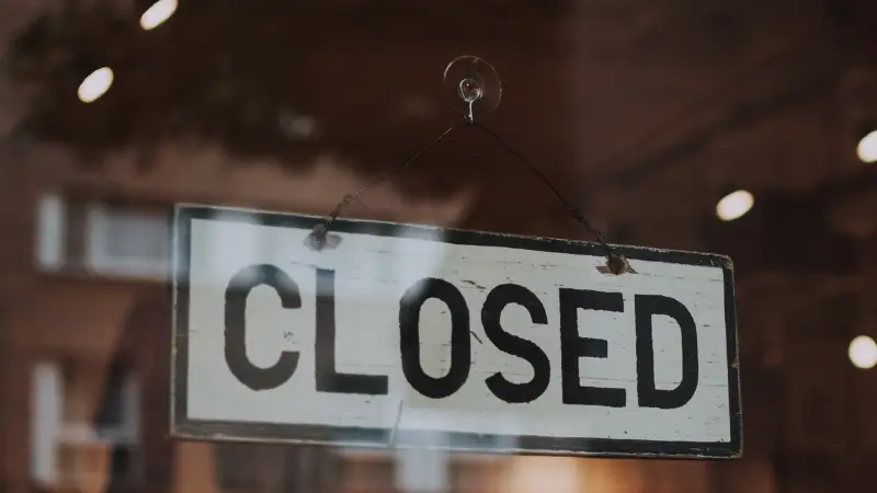 A weathered "CLOSED" sign hangs from a suction cup on a glass window, with reflections of lights and buildings visible in the background. The image conveys that the business or establishment is not currently open, symbolizing closure, pause, or downtime.