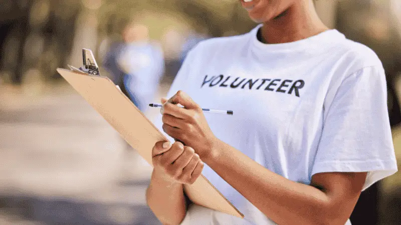 A smiling volunteer wearing a white T-shirt with the word "VOLUNTEER" printed on it is holding a clipboard and writing with a pen. The background is softly blurred, suggesting an outdoor setting with other people in the distance. The image conveys themes of community service, organization, and engagement.