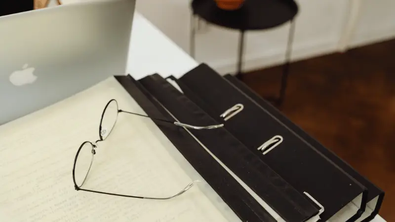 A pair of black-rimmed glasses resting on open paperwork, with a laptop and folders nearby.