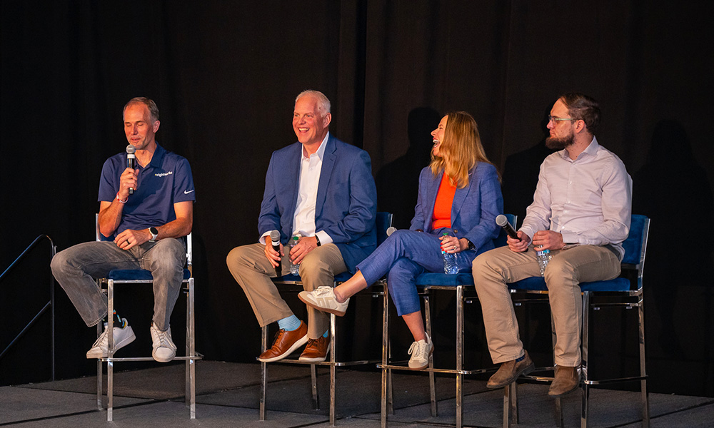Four professional panelists sitting on high stools on a stage with a dark backdrop during a technology discussion at the RightNOW26 conference. One panelist on the left is speaking into a microphone, while the others look on and smile.