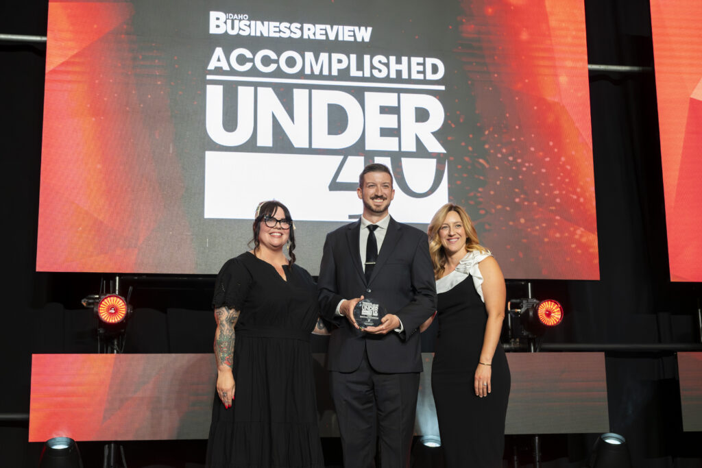 Matthew Goodfellow stands on stage holding his Accomplished Under 40 award trophy alongside two presenters at the Idaho Business Review awards gala.