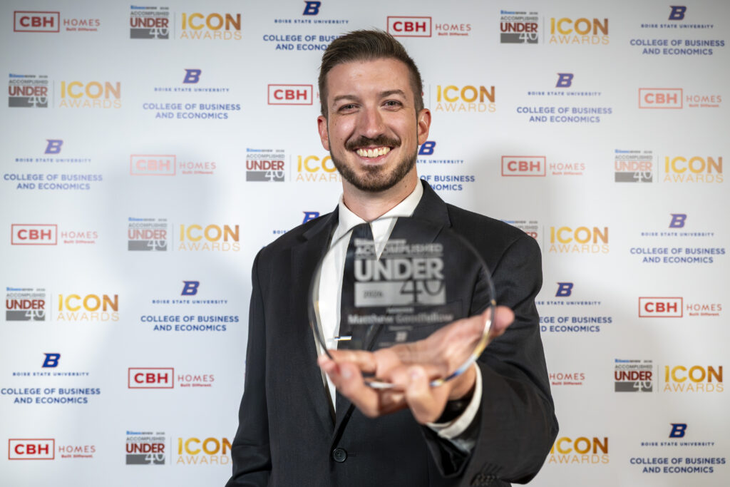 Matthew Goodfellow smiling and holding up his Idaho Business Review Accomplished Under 40 award in front of an event backdrop.